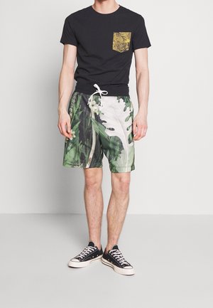 Homme debout portant un t-shirt noir avec une poche feuille jaune et un short à cordon avec un motif feuillu vert, associé à des baskets noires.