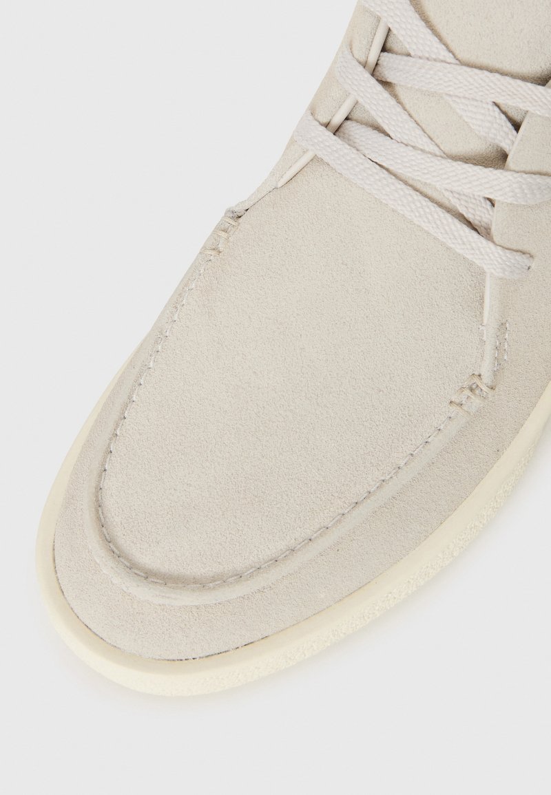 Chaussure en suede gris clair avec un embout arrondi, un dessus texturé et une semelle en caoutchouc de couleur crème. Elle présente des lacets plats et des détails de couture subtils.