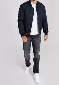 Muž v námořnické bomber bundě přes bílé polokošile, černých vybledlých džínách a bílých teniskách kráčející na jednobarevném pozadí.