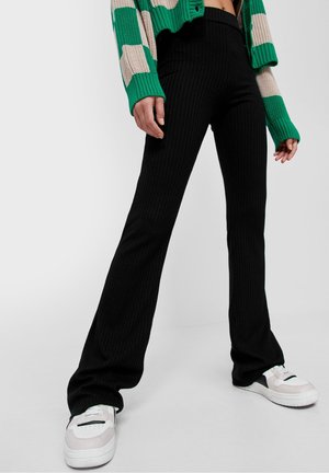 Zwarte geribbelde flared broeken met een hoge taille, gecombineerd met witte sneakers. Achtergrond is eenvoudig wit, waardoor de kleding opvalt.