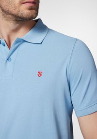 Jasnoniebieska koszulka polo z kołnierzykiem, trzema guzikami oraz małym, czerwonym haftowanym logo na lewej piersi. Teksturowana tkanina.