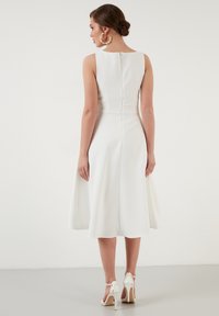 Rochie albă fără mâneci, de lungime medie, cu un corset ajustat și o fustă evazată. Spate cu deschidere în formă de cheie și detaliu de cusătură. Asortată cu sandale albe cu toc înalt.