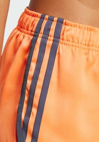 Orange athletic shorts made of smooth, lightweight fabric, featuring a gathered waistband and two vertical navy stripes on the side.