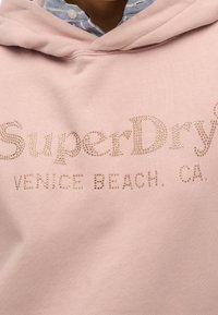 Sudadera rosa con un acabado texturizado, con letras de clavos dorados que dicen "SuperDry" y "VENICE BEACH, CA" en la parte frontal. Material de mezcla de algodón.