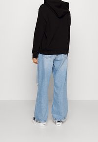 Hoodie noir à coupe ample avec capuche, porté avec un jean bleu clair à jambes larges et des chaussures blanches sans lacets.