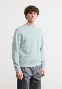 Νεαρός άνδρας με σγουρά μαλλιά, φορώντας ένα ανοιχτό μπλε φούτερ BOSS και γκρι παντελόνι, σε λευκό απλό φόντο.