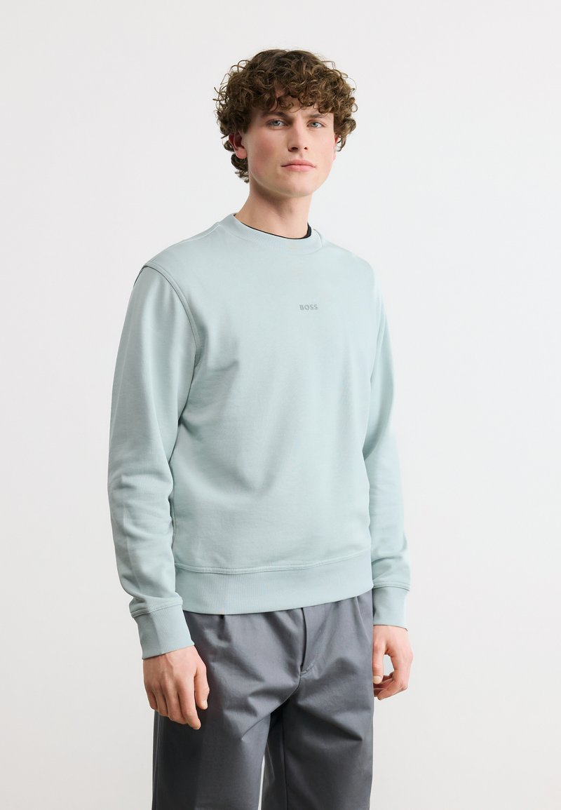 Νεαρός άνδρας με σγουρά μαλλιά, φορώντας ένα ανοιχτό μπλε φούτερ BOSS και γκρι παντελόνι, σε λευκό απλό φόντο.