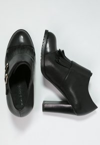 Zapatos de cuero negro con tacón, puntera redondeada, flecos decorativos y detalle de hebilla. Presentan un tacón grueso y una suela texturizada.