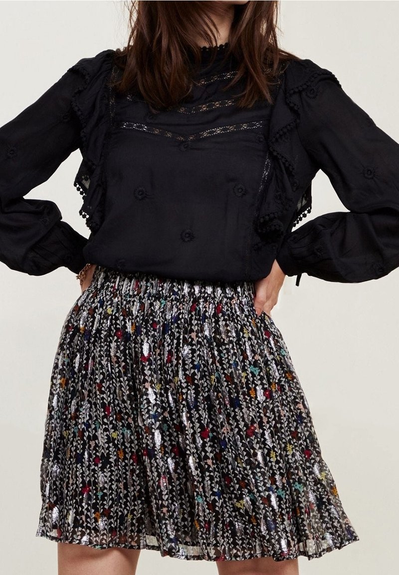Schwarze Bluse mit Spitzenakzenten und Rüschensäumen, kombiniert mit einem gemusterten Rock, der bunte Blumenmuster auf dunklem Hintergrund zeigt.