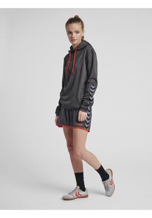 Grå atletisk hoodie med røde accenter og logo, parret med grå shorts med rød kant og chevron mønster; model iført grå og røde sko.