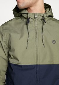 Huvjacka med tvåfärgad design, olivgrön upptill och marinblå längst ner. Texturerat tyg med justerbara dragsnören och dragkedja.