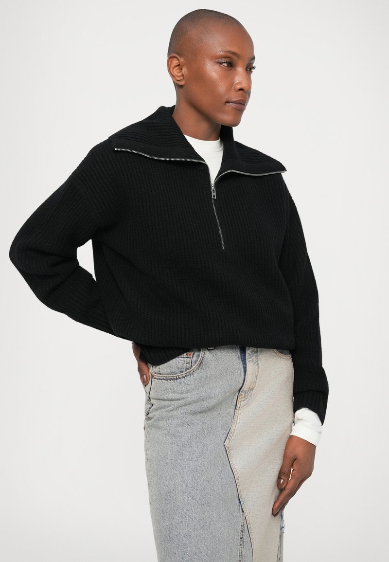 Svart ribbad tröja med hög krage och silvrig dragkedja, i kombination med en ljusblå denimkjol med kontrasterande paneler och sömmar.