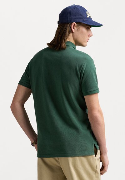 Groene poloshirt met een gestructureerde afwerking, korte mouwen en zijslits. De persoon draagt een marineblauwe pet met een geborduurd logo.
