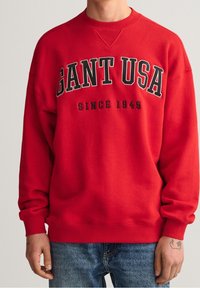 Sudadera roja con cuello redondo, que presenta un texto negro en negrita "GANT USA" y "SINCE 1949". El material parece suave, con un ajuste casual.