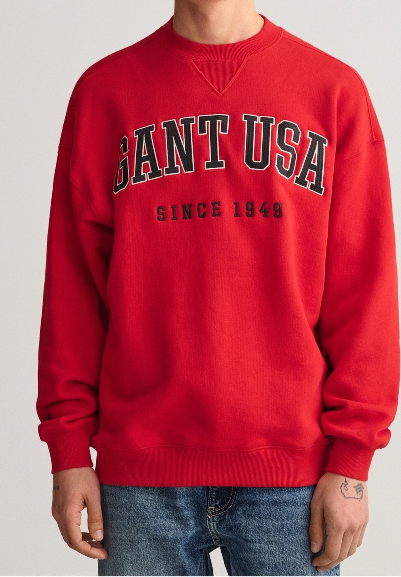 Sudadera roja con cuello redondo, que presenta un texto negro en negrita "GANT USA" y "SINCE 1949". El material parece suave, con un ajuste casual.