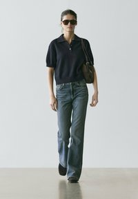 Dunkelblaues Polohemd mit kurzen Ärmeln, hoch taillierte hellblaue Jeans, schwarze Slip-On-Schuhe und eine kleine braune Tasche; große Sonnenbrillen runden den Look ab.