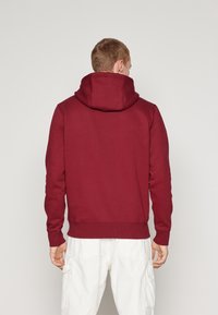 Sudadera burdeos con una gran capucha y puños acanalados, hecha de una tela suave. El diseño presenta un corte clásico con la parte trasera lisa.