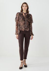 Chemisier brun transparent avec imprimé léopard, manches volantées et col à nouer, associé à un pantalon ajusté brun foncé et des escarpins noirs à lanière T.