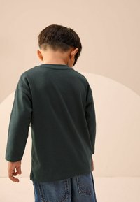 Koszula z długim rękawem w głębokim zielonym kolorze z bawełny, z okrągłym dekoltem i luźnym krojem, w zestawie z niebieskimi szortami dżinsowymi z tylnymi kieszeniami.