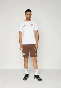 Muž stojící v bílém tričku Puma a hnědých šortkách Puma se sportovními logy, v bílých ponožkách a černých teniskách na jednobarevném pozadí.