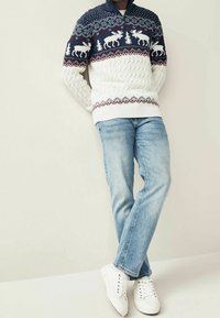 Pull en tricot bleu marine et blanc, avec un motif en point de câble représentant des orignaux et des arbres, associé à un jean bleu clair et des baskets blanches.