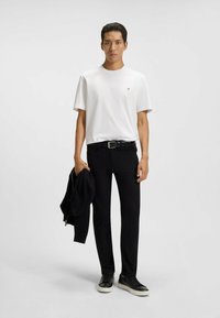 T-shirt bianca a maniche corte con un piccolo logo, abbinata a pantaloni neri attillati e sneakers neri. Il modello tiene in mano una giacca nera.
