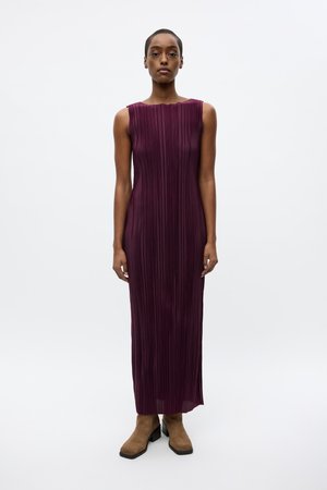 Plum-colored, floor-length pleated dress with a scalloped neckline and sleeveless design, paired with tan ankle boots on a plain background.