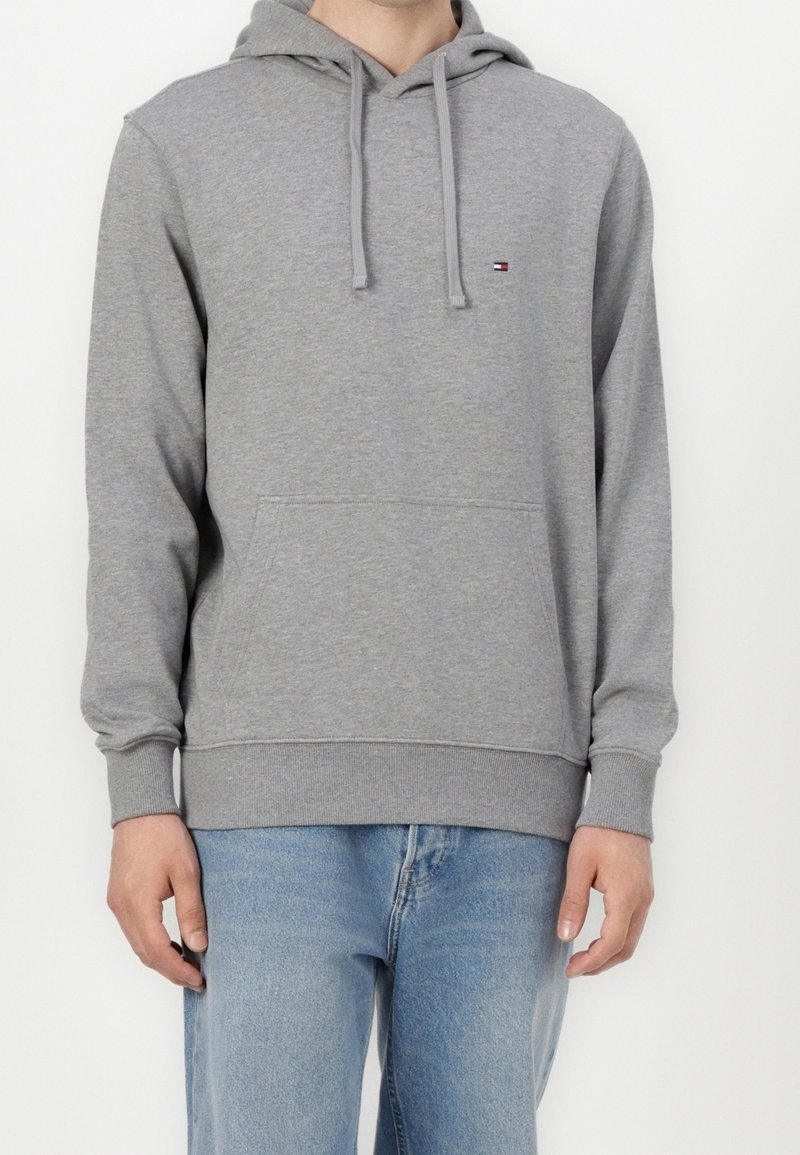 Personne portant un sweat à capuche gris uni avec cordons et un petit logo sur la poitrine, assorti à un jean bleu clair sur fond blanc.