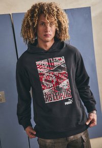 Sort hættebluse med et grafisk print, der indeholder rød, hvid og sort tekst, geometriske mønstre og PUMA-logoet nederst.