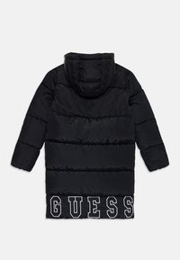 Fekete puffermellény kapucnival, függőleges steppelt mintázattal és nagy fehér "GUESS" logóval az alján. Simább, fényes anyag.
