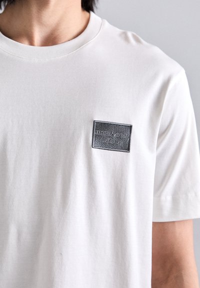 White cotton t-shirt featuring a rectangular gray leather patch with embossed logo on the left chest, crew neckline, and short sleeves.