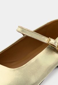 Detailný záber na kovovú zlatú topánku Mary Jane s tenkým remienkom a zlatou prackou na béžovej vnútornej podrážke.