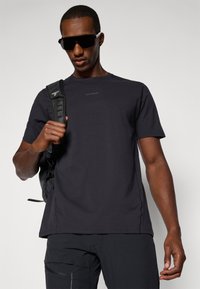 Tricou negru cu mânecă scurtă, cu guler rotund și material texturat, purtat cu un rucsac negru și ochelari de soare închiși la culoare.