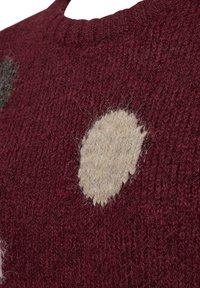 Närbild av en vinröd stickad tröja med en beige oval applikation och texturerade ullfibrer synliga på tygytan.