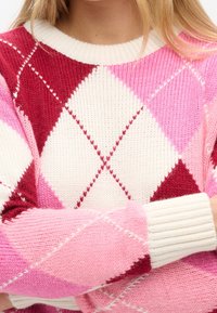 Donna che indossa un maglione lavorato a maglia a rombi nei colori rosa, rosso e bianco, con le braccia incrociate, esibendo il tessuto testurizzato e il colletto e i polsini a coste.