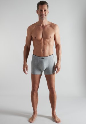 Adult man standing barefoot, shirtless, wearing fitted gray boxer briefs against a plain light background.