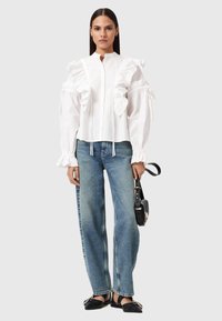 Kvinna i en vit volangblus, blå jeans, svarta spetsiga ballerinaskor, och håller en svart handväska mot en enfärgad ljus bakgrund.