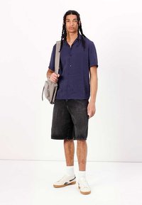 Marineblå kortærmet skjorte med knapper, sorte denimshorts, hvide sneakers med tan sål, beige messenger-taske, stående foran en ensfarvet baggrund.