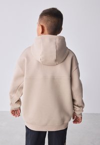 Smėlinės spalvos džemperis su kapišonu, tekstūruota nugara, ribinėmis rankogaliais, laisvo silueto ir su pamuštu kapišonu; dėvimas vaiko iš nugaros.