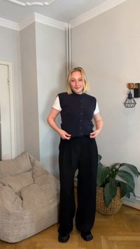 Marineblå vest med knapper og høj krave, båret over en hvid t-shirt, kombineret med brede sorte bukser og sorte sko. Indendørs miljø med planter.