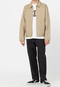 Man som bär en beige zip-up jacka, vit t-shirt med grafik, svarta byxor och svart-vita sneakers, står mot en vit bakgrund.