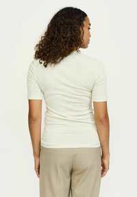 Gaiši bēšs, īsām piedurknēm tops no mīksta auduma, pieguļošs dizains, ar apaļu kakla izgriezumu un gludu tekstūru, valkāts kopā ar bēšām biksēm.