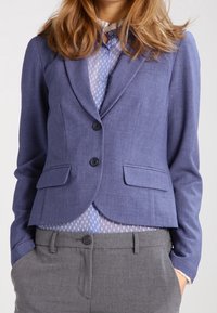 Jasnoniebieska skrojona marynarka z dwiema guzikami, kieszeniami z klapkami i dopasowanym fasonem, noszona na jasno-niebieskiej bluzce w kropki i szarych spodniach.