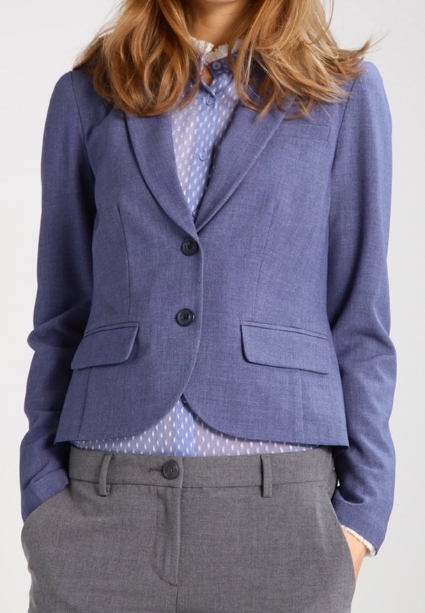 Jasnoniebieska skrojona marynarka z dwiema guzikami, kieszeniami z klapkami i dopasowanym fasonem, noszona na jasno-niebieskiej bluzce w kropki i szarych spodniach.
