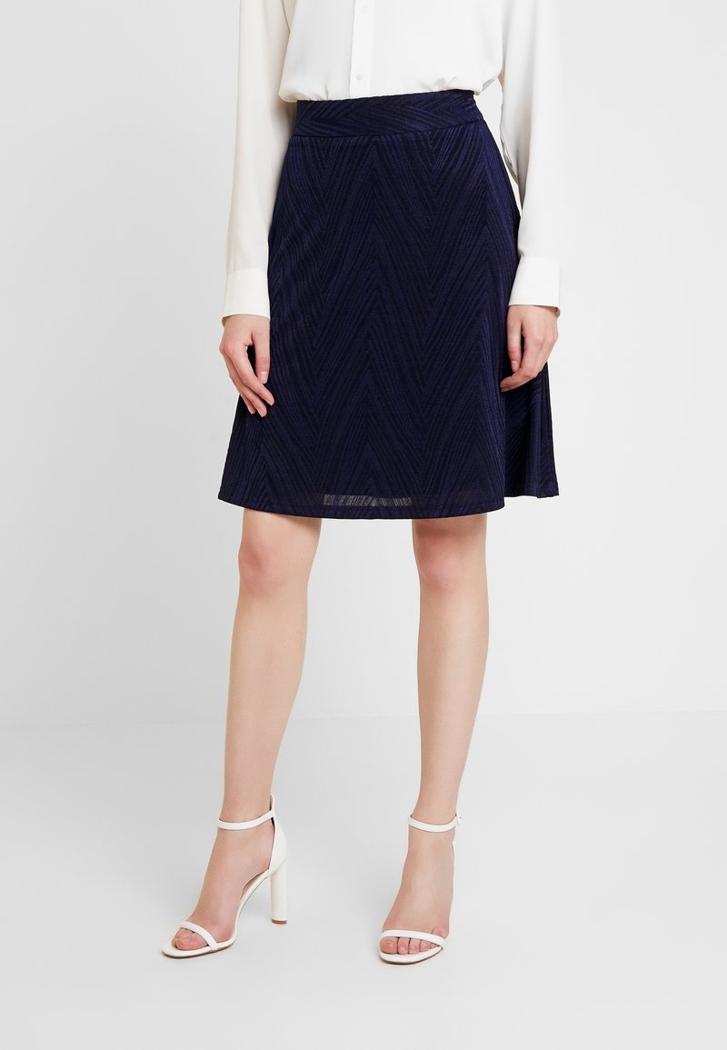 Ein marineblauer Rock mit Chevron-Muster, der eine taillierte Taille und einen ausgestellten Saum hat. Das Outfit wird mit weißen, schnittigen High-Heels kombiniert.