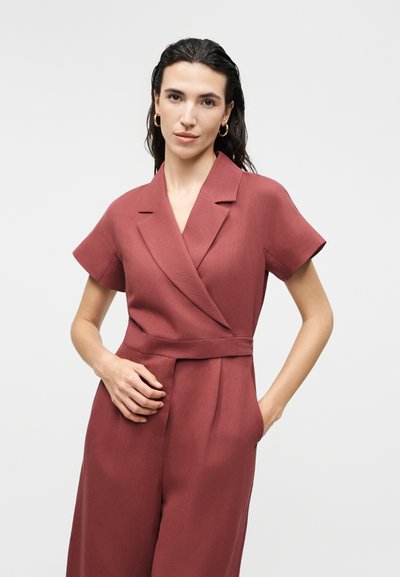 Γυναίκα με σκούρα μαλλιά φοράει ρούχο ολόσωμο με κοντά μανίκια σε σκουρόχρωμη σκουριά, με γιακά και ζώνη στη μέση, στέκεται μπροστά σε απλό φωτεινό φόντο.