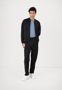 Zwarte sportjack met gestreepte mouwen, gecombineerd met een blauwe crewneck en zwarte broek. Model draagt witte sneakers. Glad materiaal, minimalistisch ontwerp.