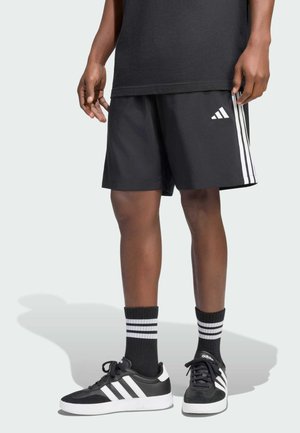 Person trägt schwarze Adidas-Shorts, schwarze Socken mit weißen Streifen und schwarze Adidas-Sneaker mit weißen Sohlen.
