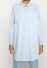 Person klädd i en ljusblå långärmad skjorta som är uppknäppt över ljusblå jeans, stående mot en enfärgad vit bakgrund.