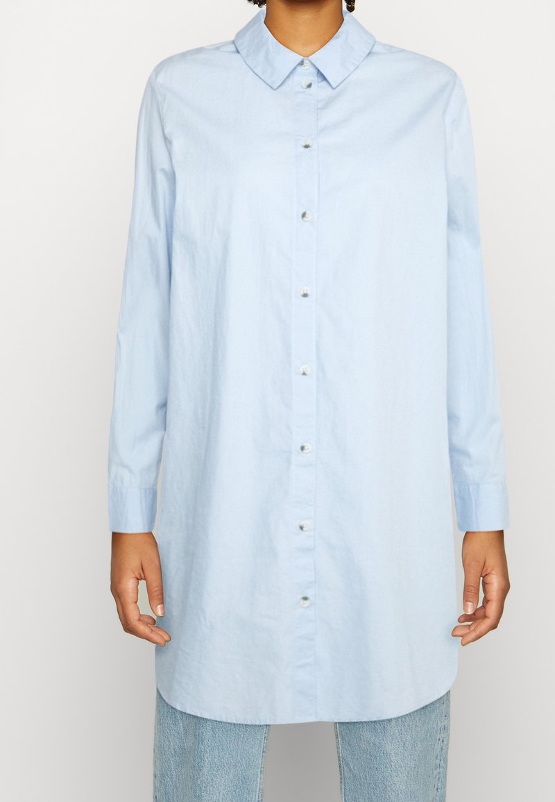 Person klädd i en ljusblå långärmad skjorta som är uppknäppt över ljusblå jeans, stående mot en enfärgad vit bakgrund.